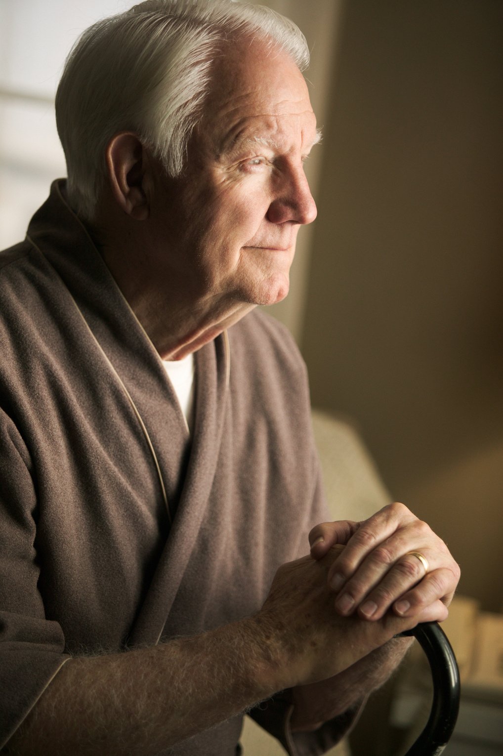 an elderly caucasian man in a brown robe sits with his cane in his bedroom